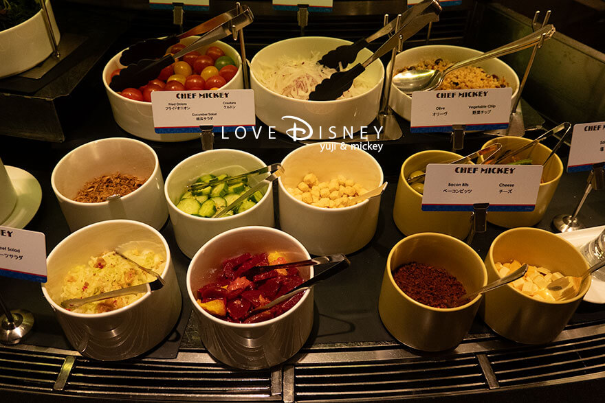 ディズニーアンバサダーホテル、シェフ・ミッキー「ディズニー・クリスマス2025ディナーブッフェ」冷たい料理（チェリートマト/オニオン/レンズ豆ともち麦のタブレ/フライドオニオン/胡瓜サラダ/クルトン/オリーブ/野菜チップ/コールスロー/ピーツサラダ/ベーコンビッツ/チーズ）